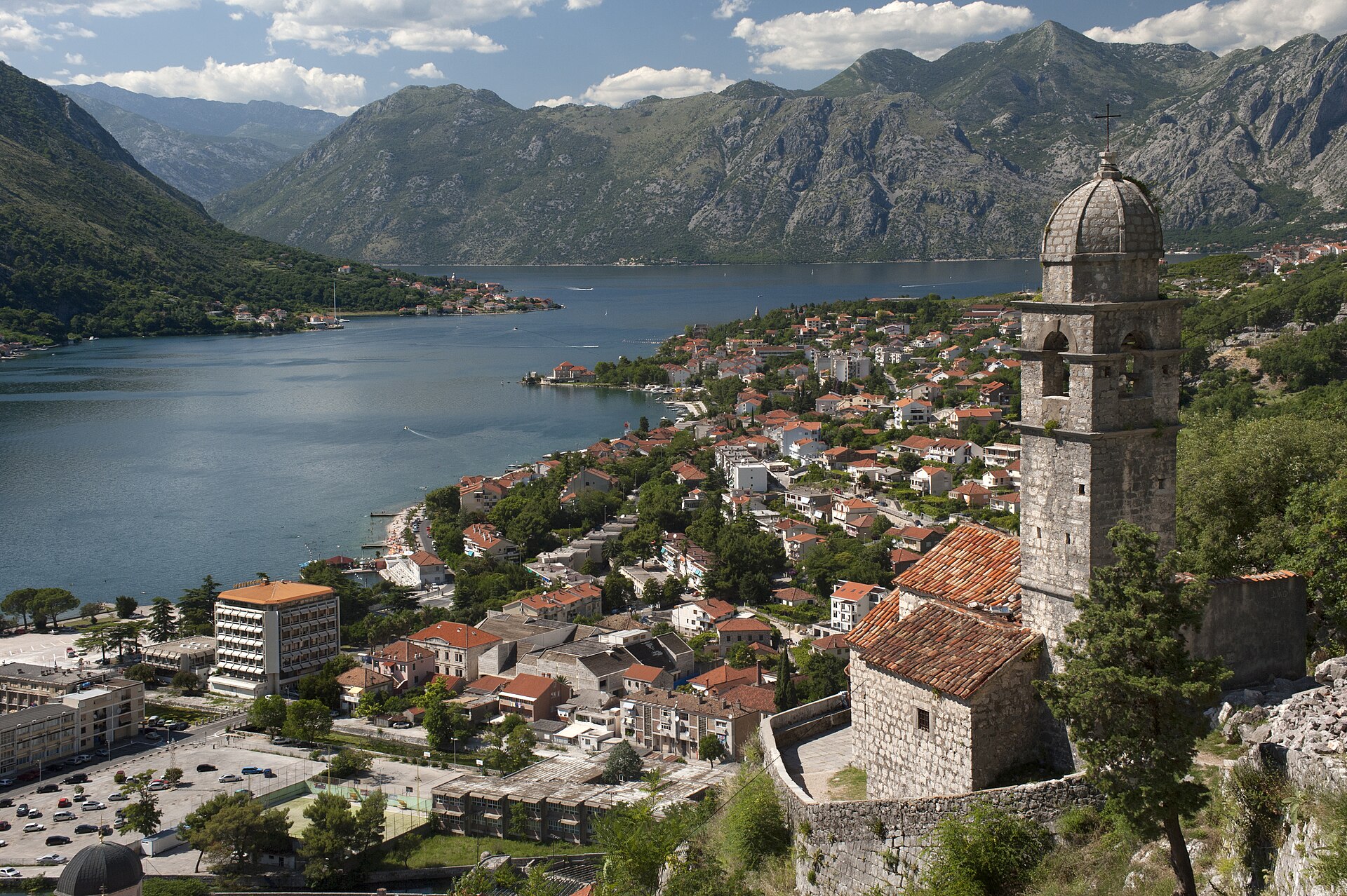 A stone church tower overlooks Kotor’s red-roofed old town and the bay, with steep mountains beyond.